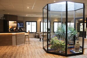Un área de bar moderna con suelo de madera, un recinto de cristal con una planta y objetos decorativos, y una pantalla de televisión.