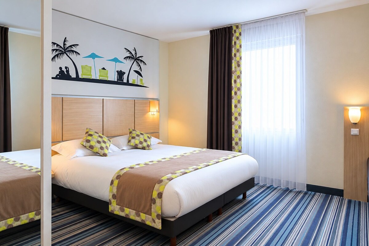 A hotel room with a large bed, a headboard with a light fixture, a wall mural, a window with curtains, and a striped carpet.