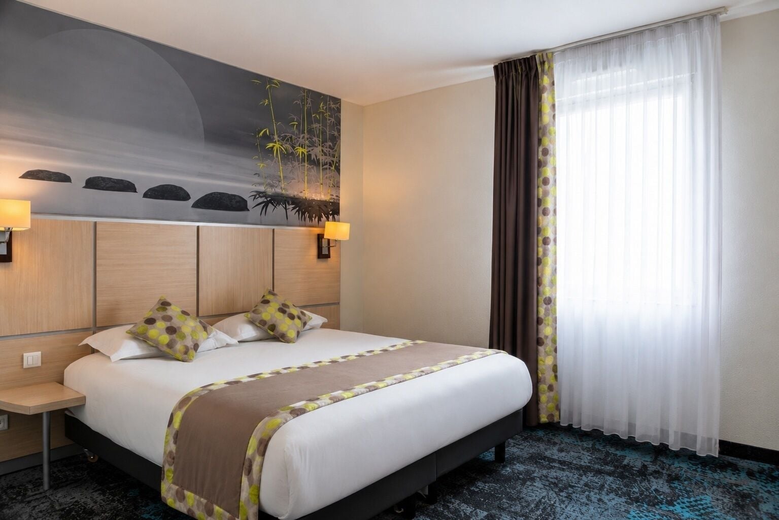 A hotel room with a bed, bedside tables, a wall-mounted lamp, a large window with curtains, and a wall art piece.