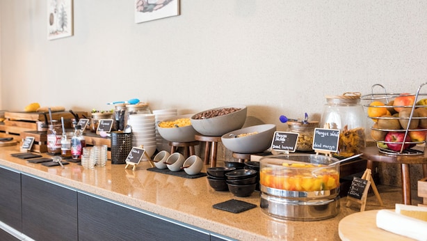 A buffet counter with various food items and condiments, including bowls of cereal, fruits, and a container of mixed fruit salad.
