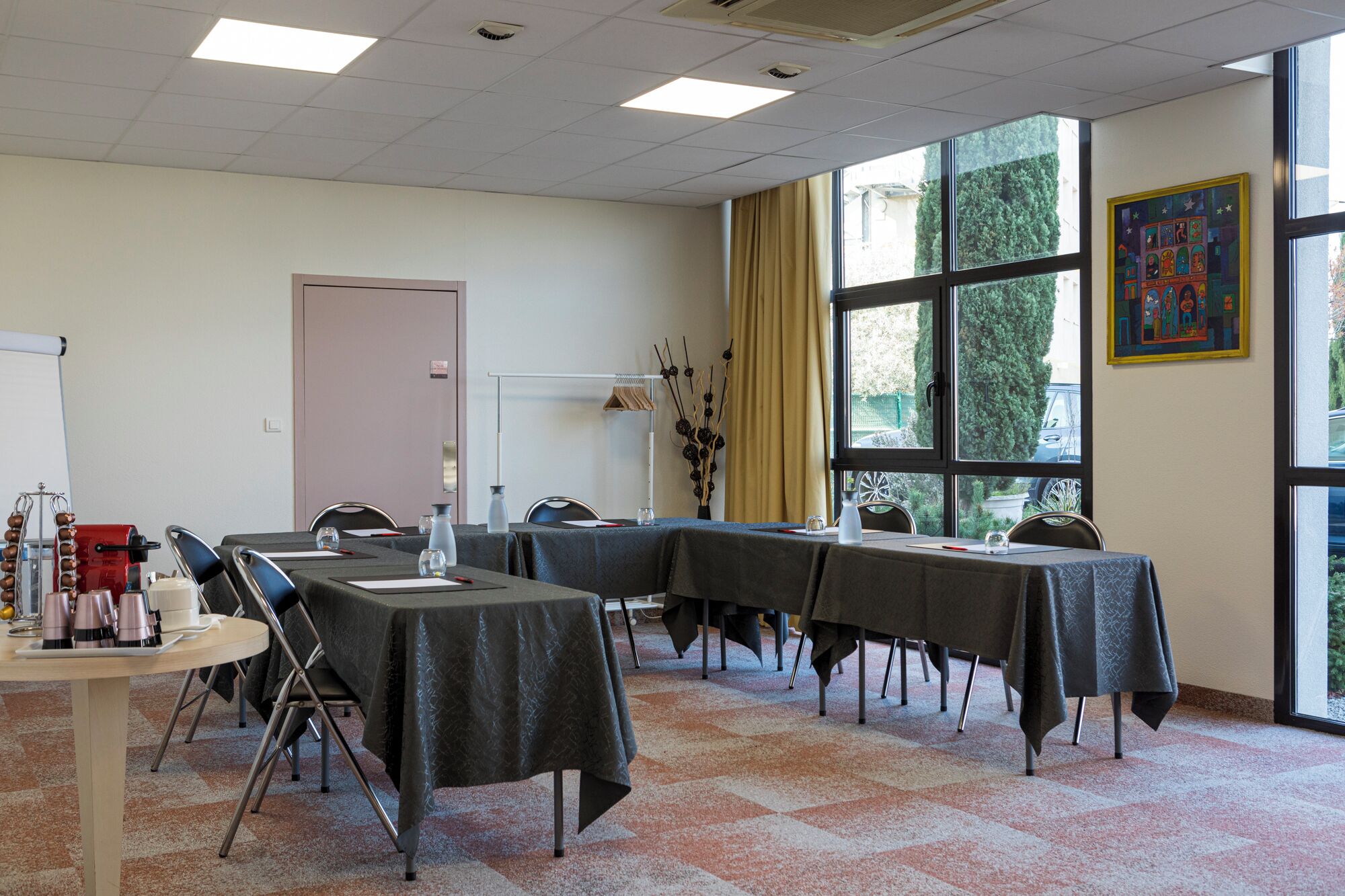 A conference room with tables, chairs, a projector, and a large window with a view of trees.