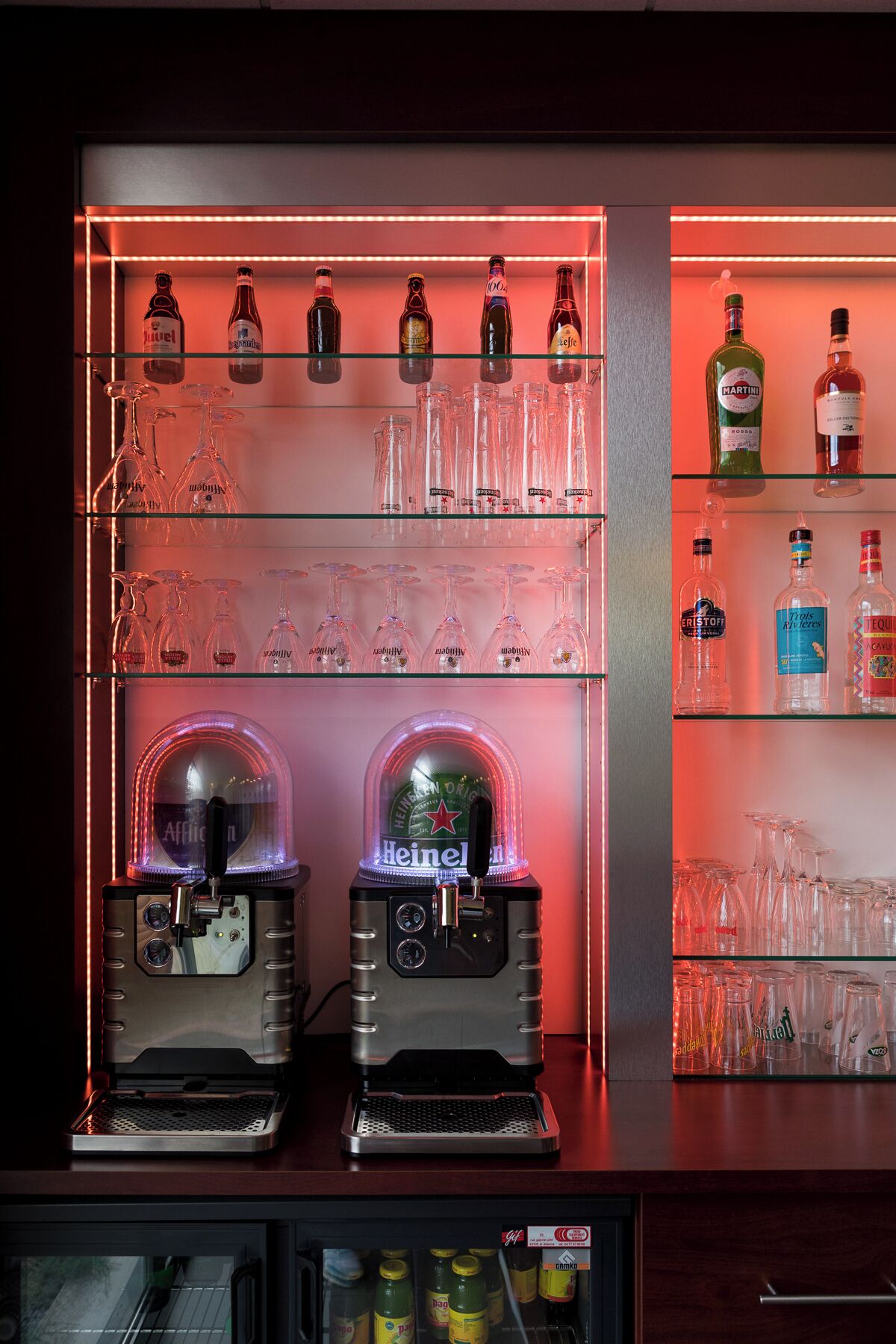 Una zona de bar con dos máquinas de café espresso, estantes llenos de botellas de licor y un refrigerador de exhibición.