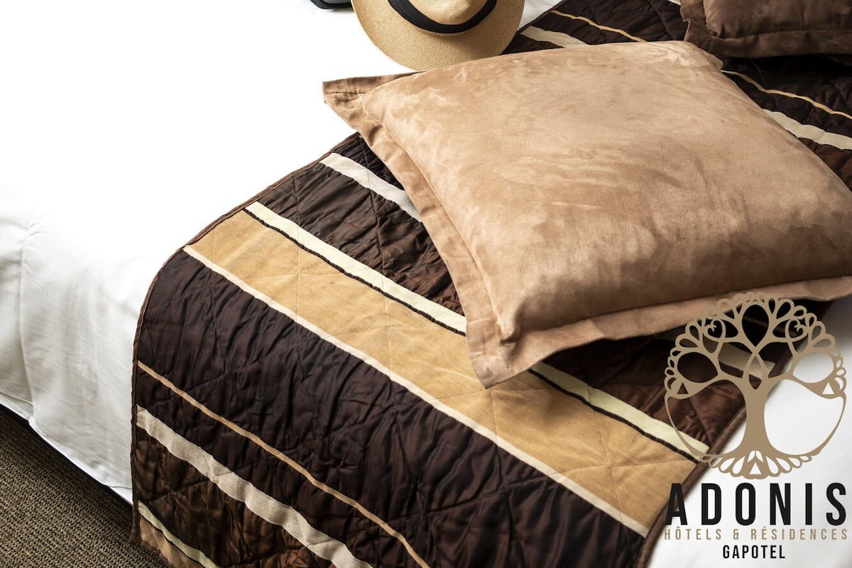 Una habitación de hotel con una cama, una maleta negra, un sombrero de paja y una almohada marrón.