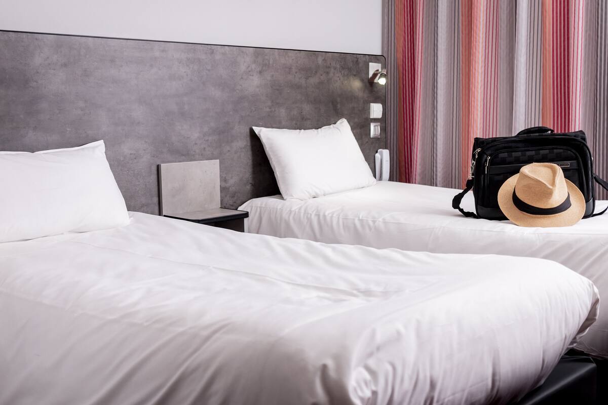 Habitación de hotel con dos camas, un cabecero gris, una cortina a rayas y un bolso negro con un sombrero.