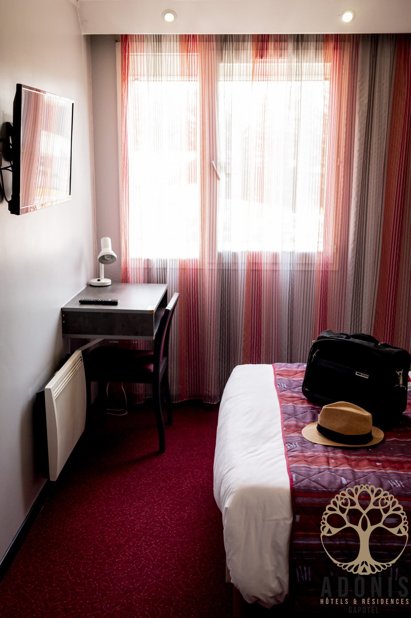 A hotel room with a bed, a desk with a lamp, a suitcase, and a hat on the bed.