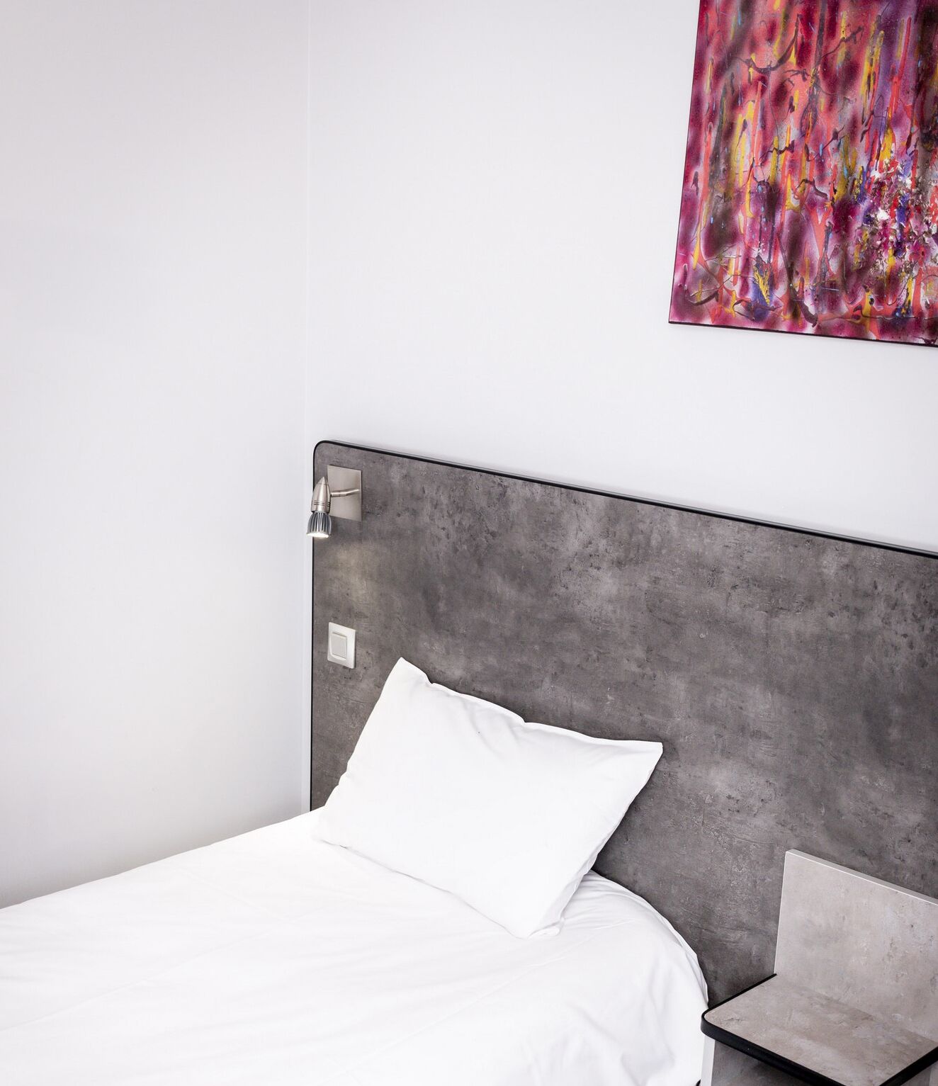 A minimalist bedroom with a grey concrete headboard, a white bedspread, and a single white pillow.