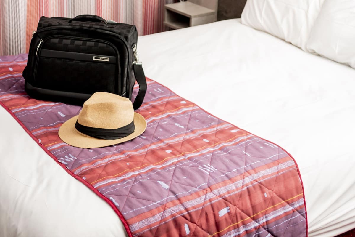 Una habitación de hotel con una cama, una maleta, un sombrero, un cuadro y una cortina.