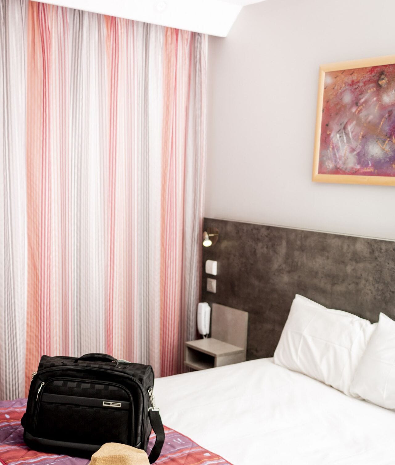 A hotel room with a bed, a nightstand, a lamp, a painting, and a black bag on the bed.