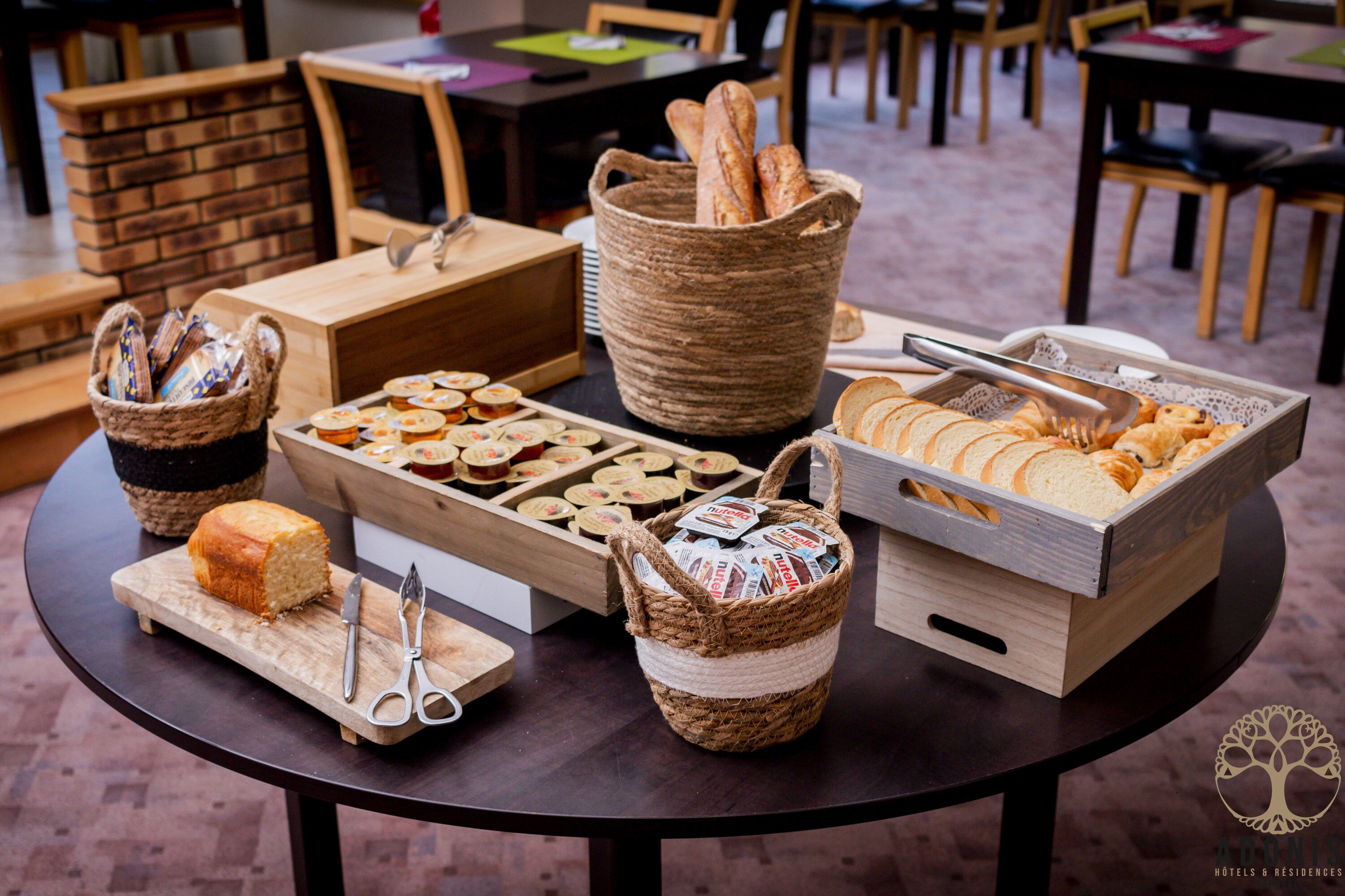 A table with various food items and utensils, including bread, pastries, and a basket of magazines.