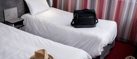 Una habitación de hotel con dos camas, una mesita de noche pequeña, una bolsa negra sobre una cama y un sombrero en el suelo.