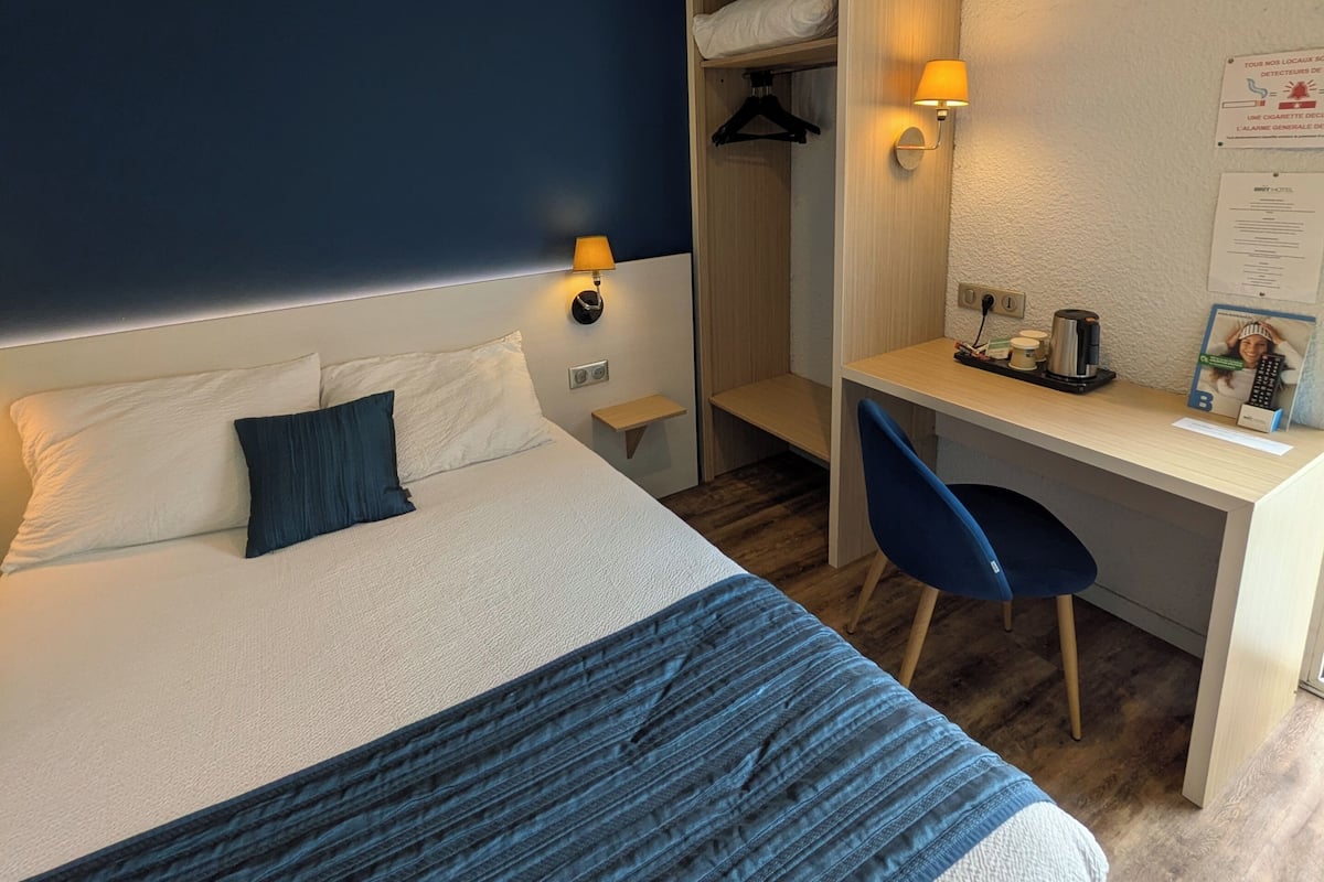 A hotel room with a bed, bedside lamps, a desk, a chair, a wardrobe, and a wall-mounted sign.