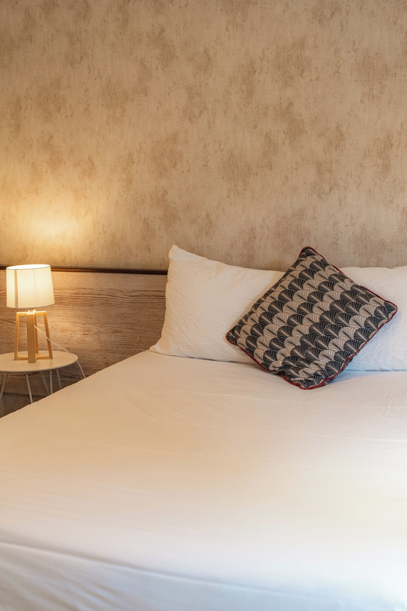 A neatly made bed with a white pillow and a patterned pillow, a bedside lamp, and a textured wall.
