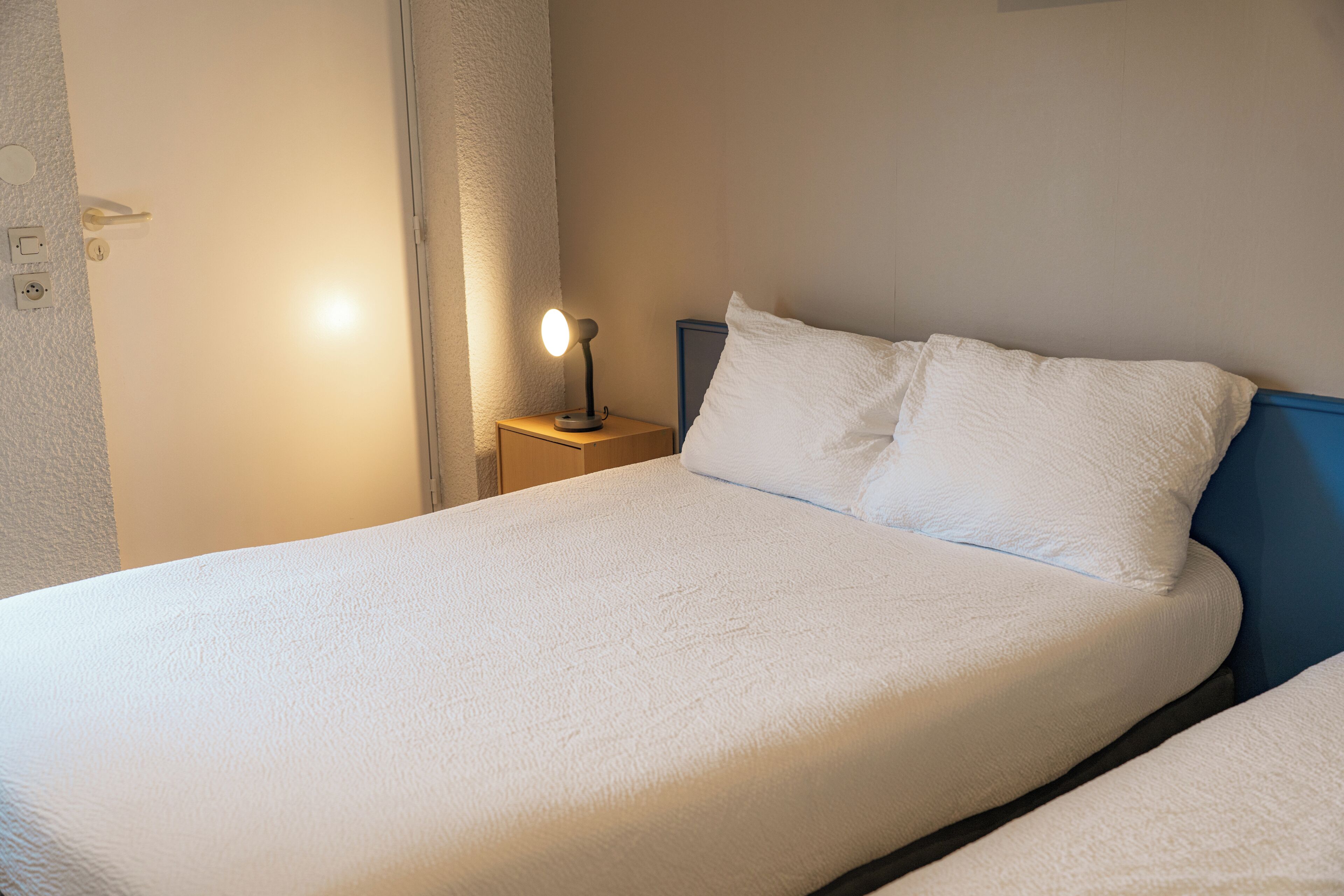 Una cama bien hecha con sábanas blancas, una lámpara de noche y una mesita de noche con toma de corriente.