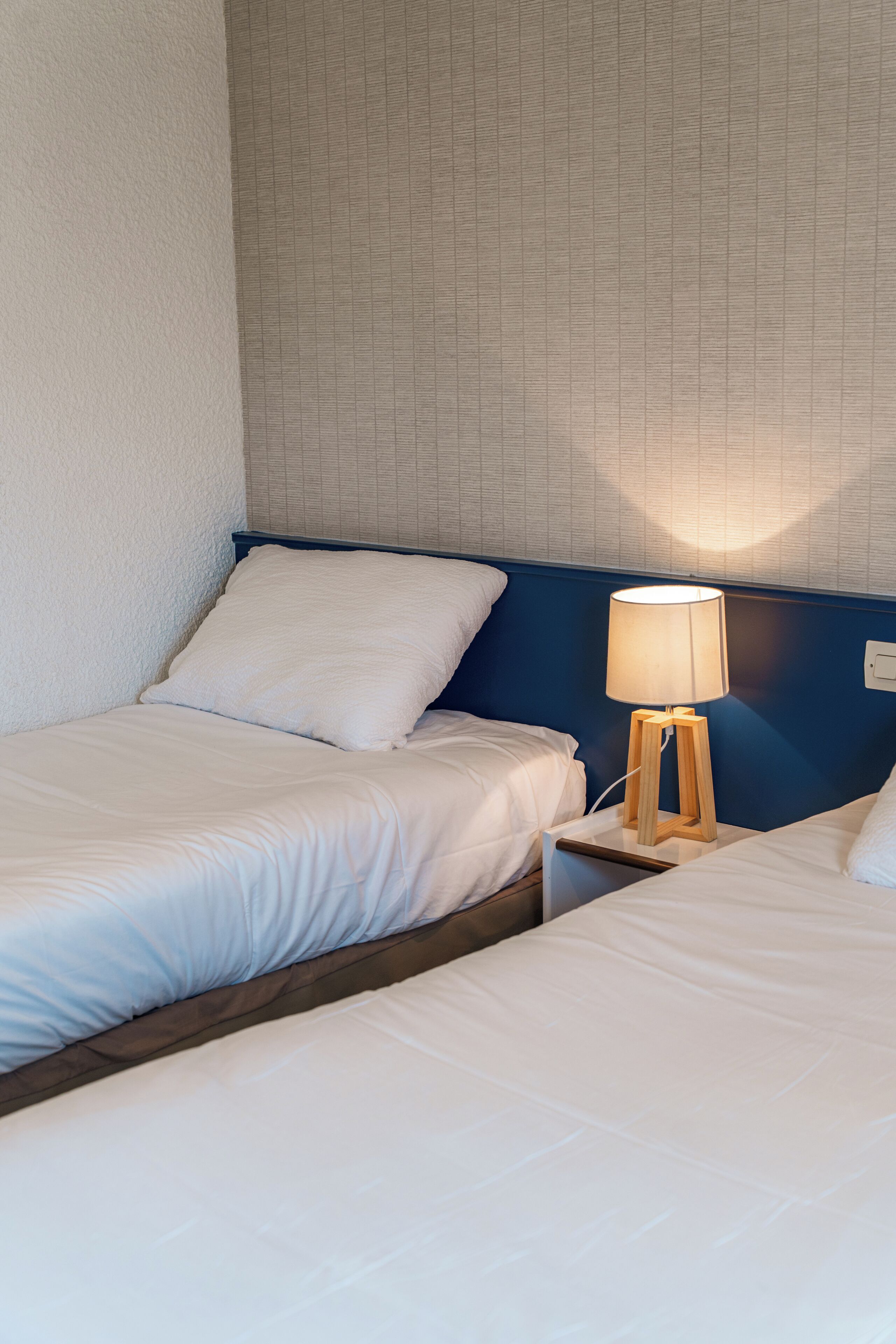 Habitación de hotel con dos camas, una mesita de noche con lámpara y pared con textura.