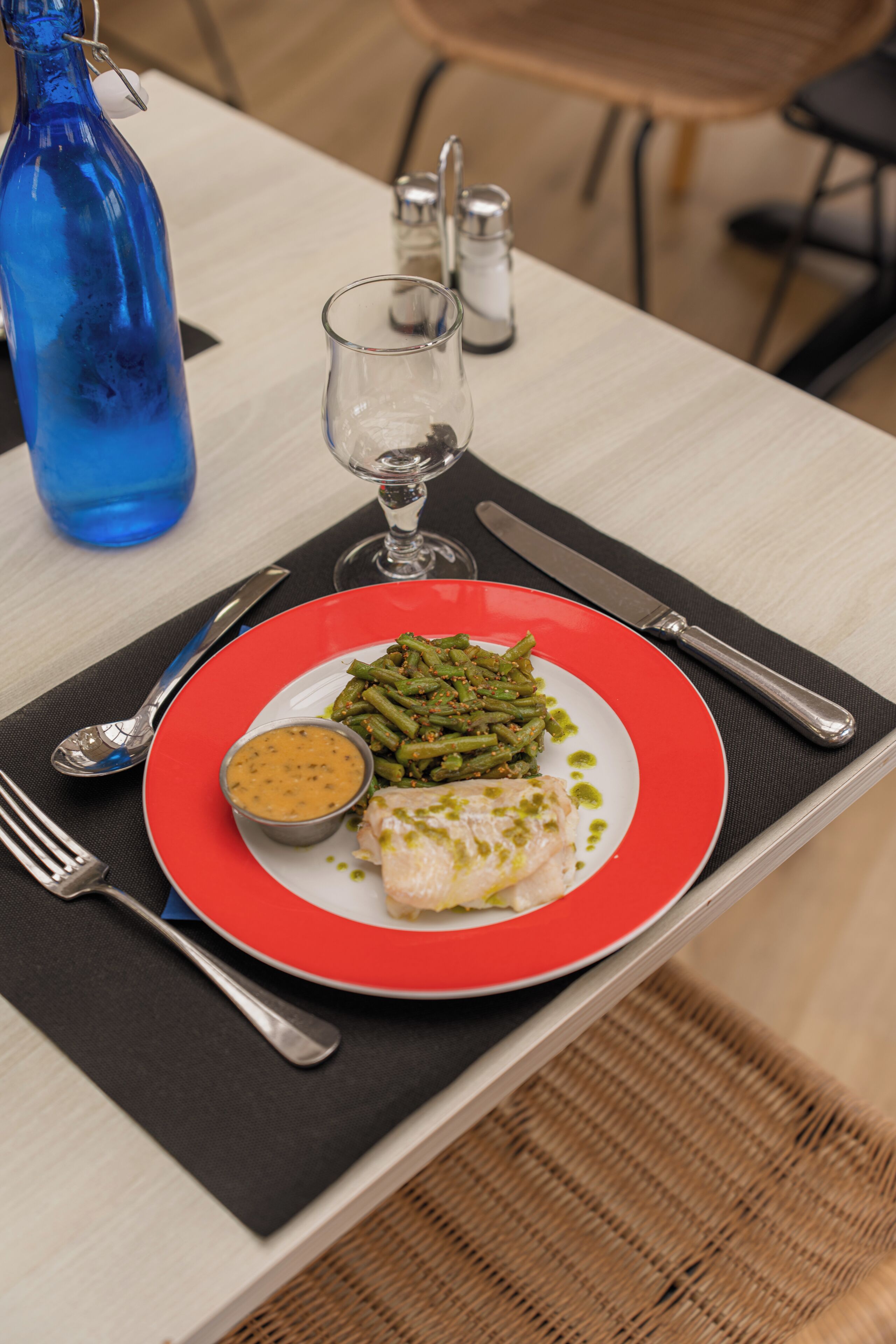 A plate of food with green beans, a piece of fish, and a sauce, set on a table with a blue bottle and a glass.