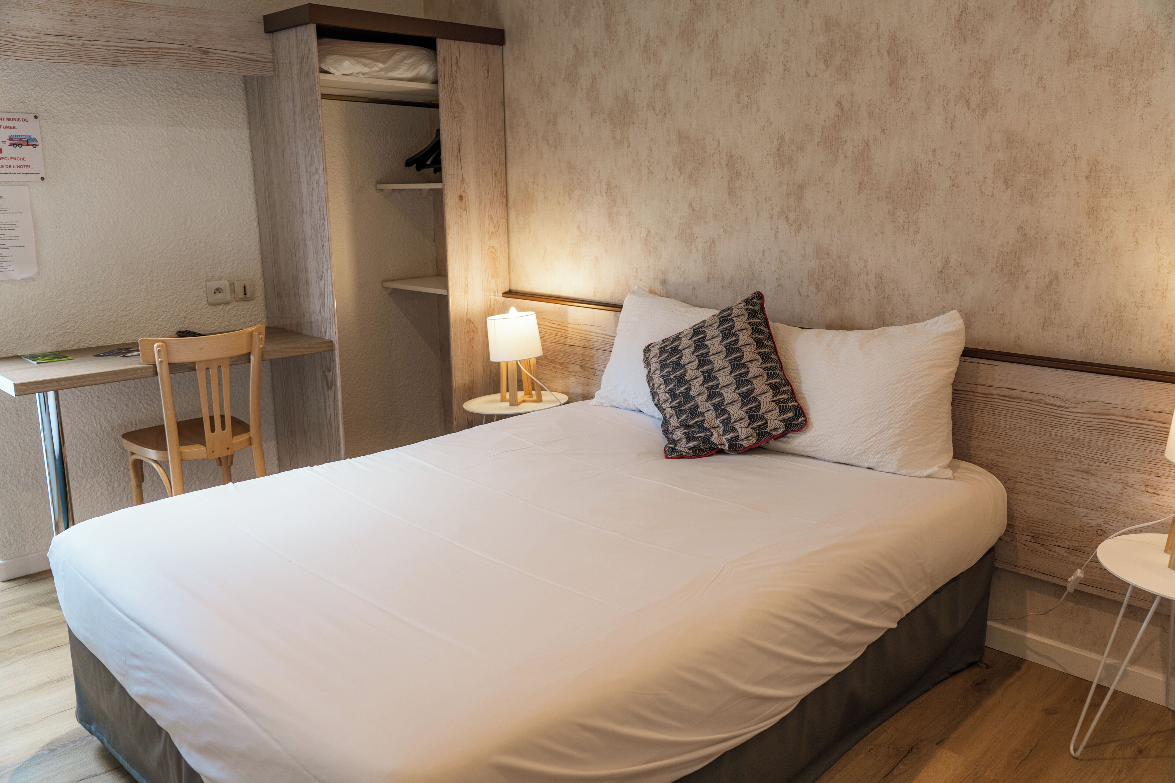 A neatly made bed with white bedding, a wooden chair, a bedside table with a lamp, and a shelf with items.