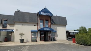 Een hotel met een bord waarop 'BRIT HOTEL' en 'ACCUEIL' staat, wat het receptiegebied aangeeft. Er is een bord voor 'ACCÈS CHAMBERS' dat naar de ingang wijst.
