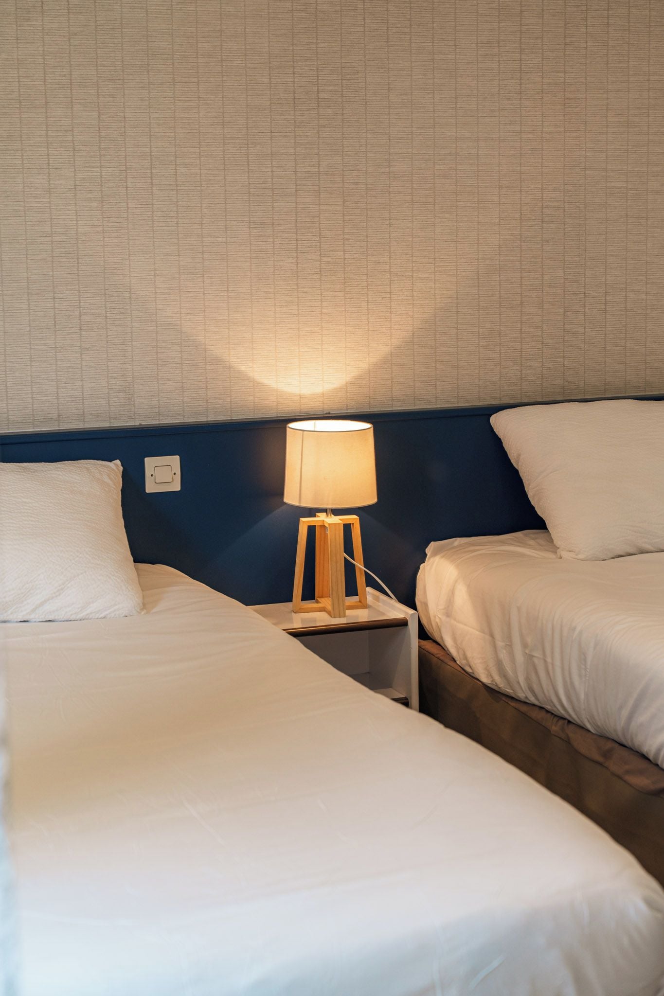 A hotel room with two beds, a bedside table with a lamp, and a wall with a textured pattern.
