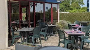 Een terras buiten met tafels en stoelen, een overdekte zithoek en een haag op de achtergrond.