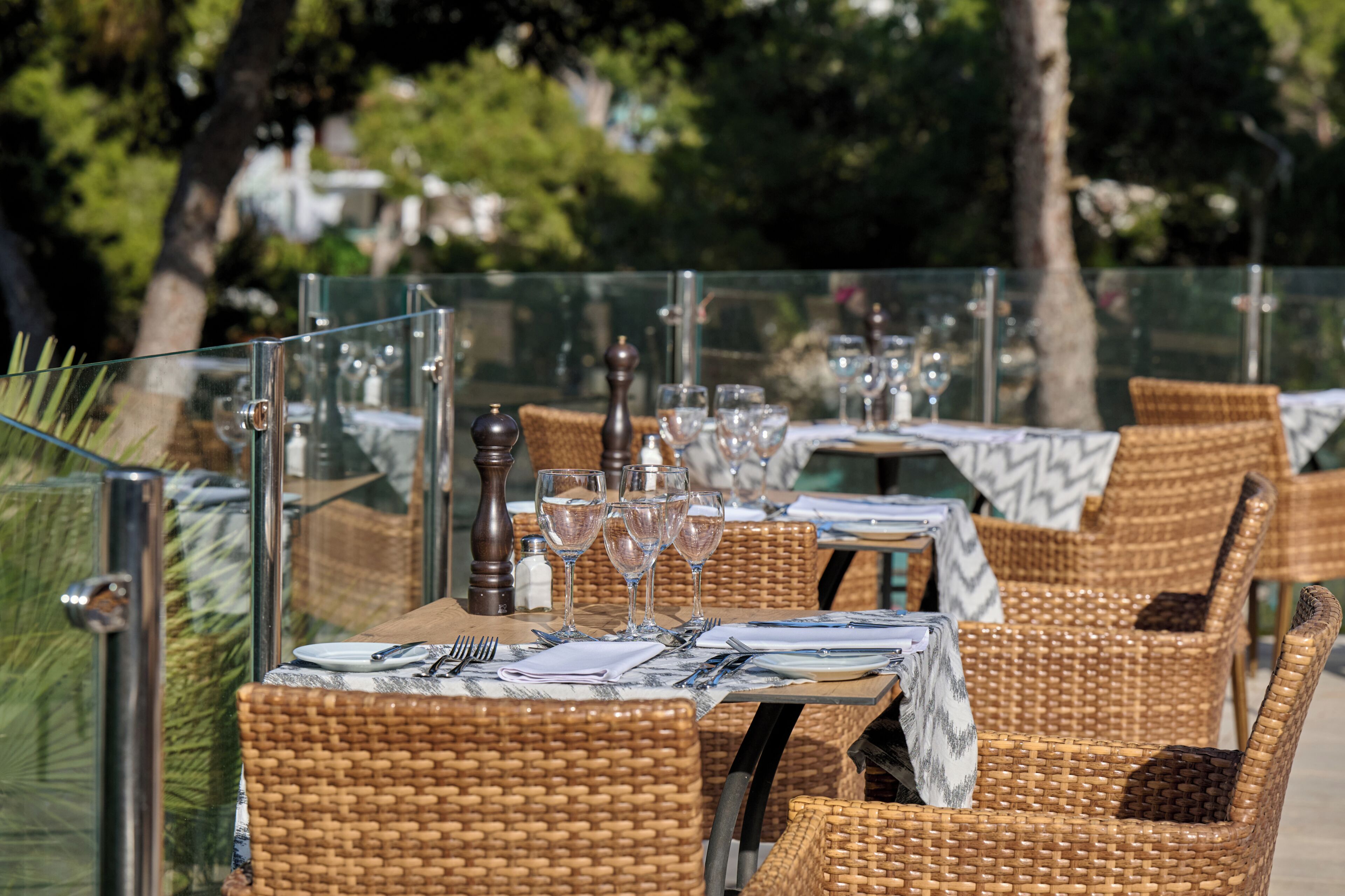 Un espace de restauration en plein air, avec des chaises et des tables en osier, chaque table étant dressée avec des couverts, des assiettes et des verres à vin.