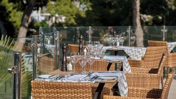 An outdoor dining area with wicker chairs and tables, each set with cutlery, plates, and wine glasses.