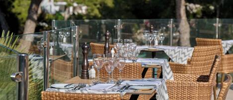 An outdoor dining area with wicker chairs and tables, each set with cutlery, plates, and wine glasses.