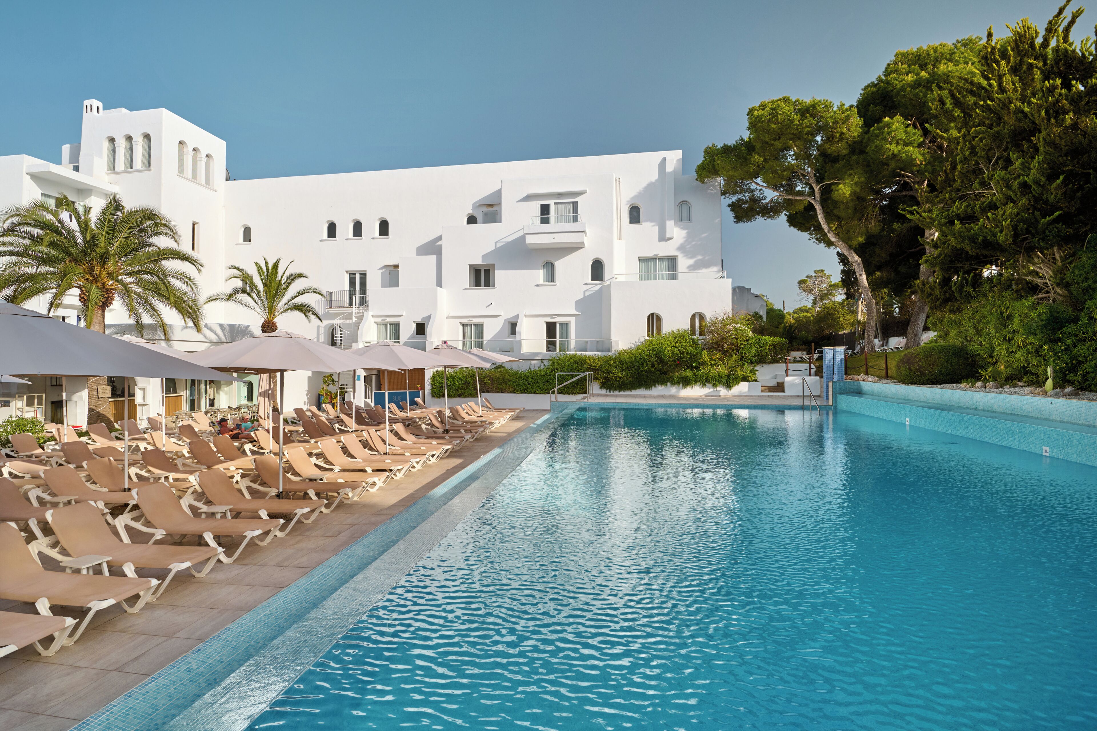 A poolside area with lounge chairs and umbrellas, a clear blue pool, and a white building with balconies.