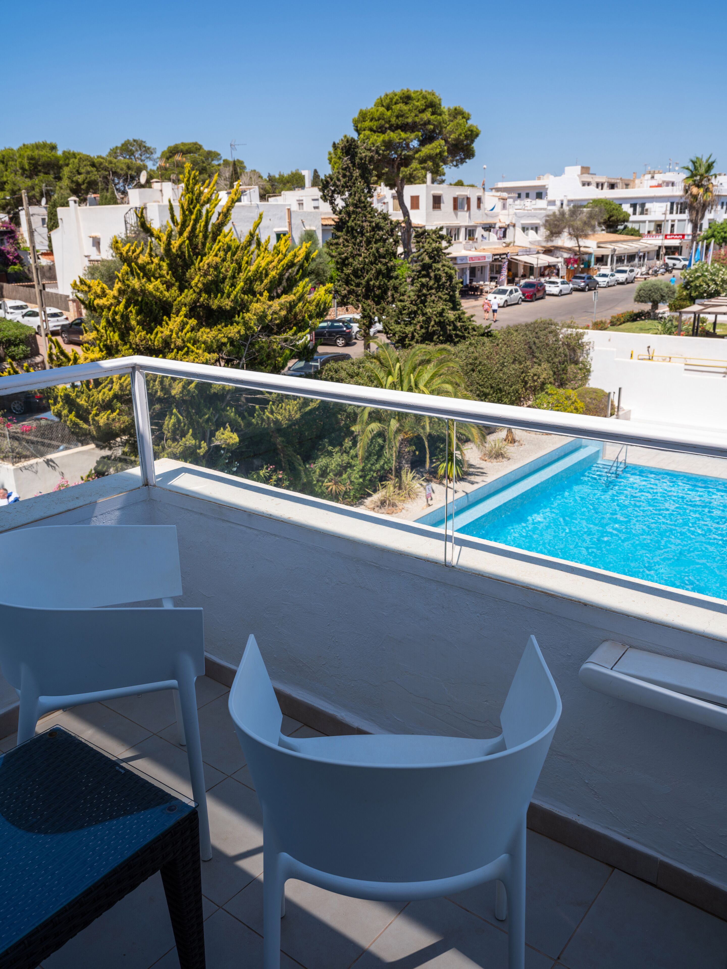 En balkon med udsigt til en pool, hvide stole og en klar blå himmel.