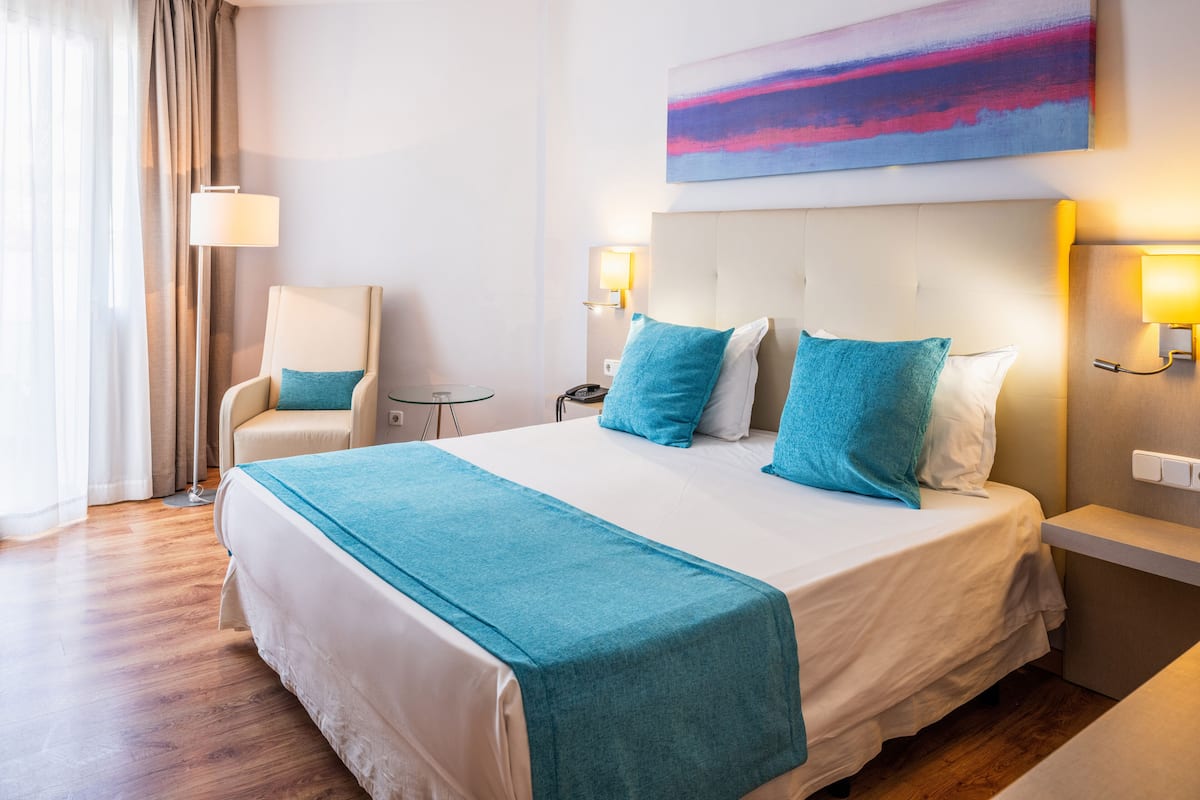Una habitación de hotel con una cama grande, una silla, una mesita y un cuadro en la pared.