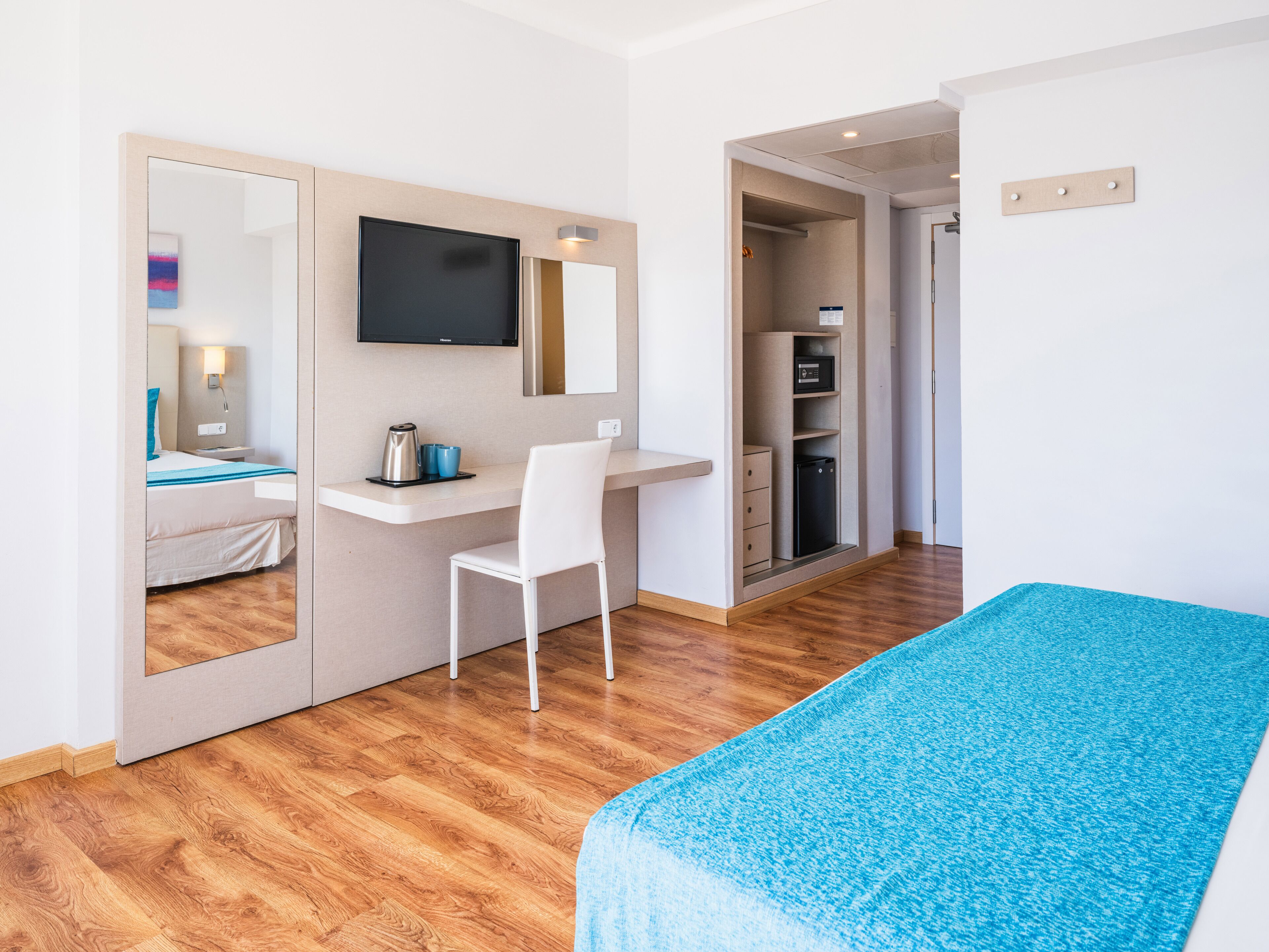 Ein modernes Hotelzimmer mit Holzboden, einem Bett mit blauer Bettdecke, einem weißen Schreibtisch mit Stuhl, einem Flachbildfernseher und einem Kleiderschrank.
