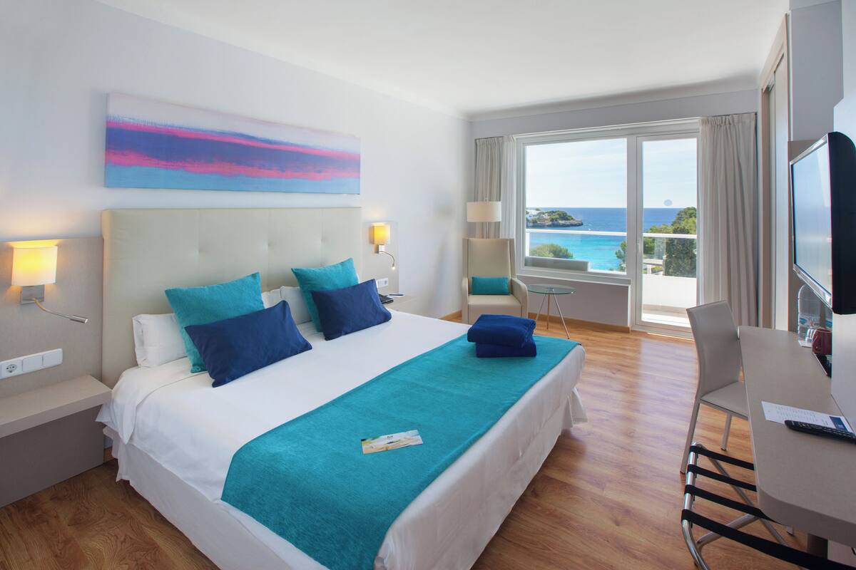 Habitación de hotel con cama, escritorio, silla, televisor y balcón con vista al mar.