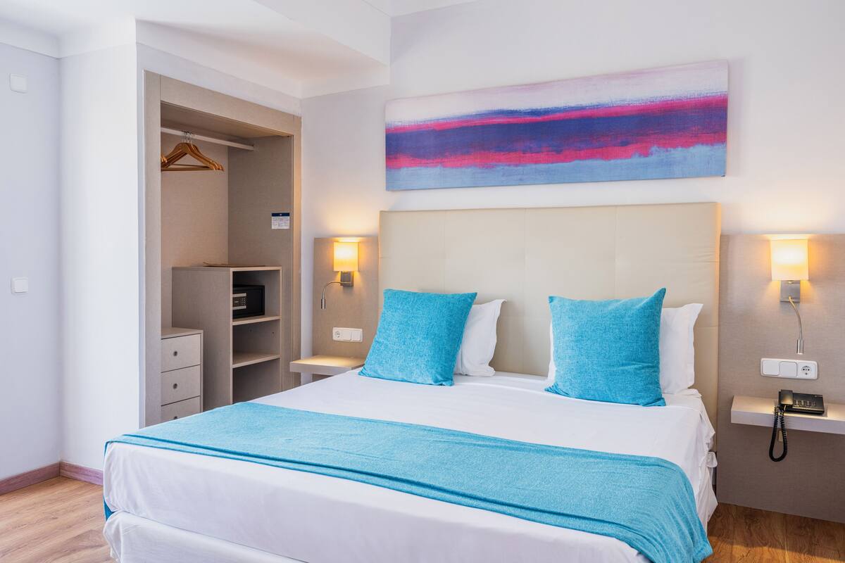 Habitación de hotel con una cama grande, dos lámparas de noche, una mesita de noche con un teléfono, un cuadro colgado en la pared y un armario con perchero.