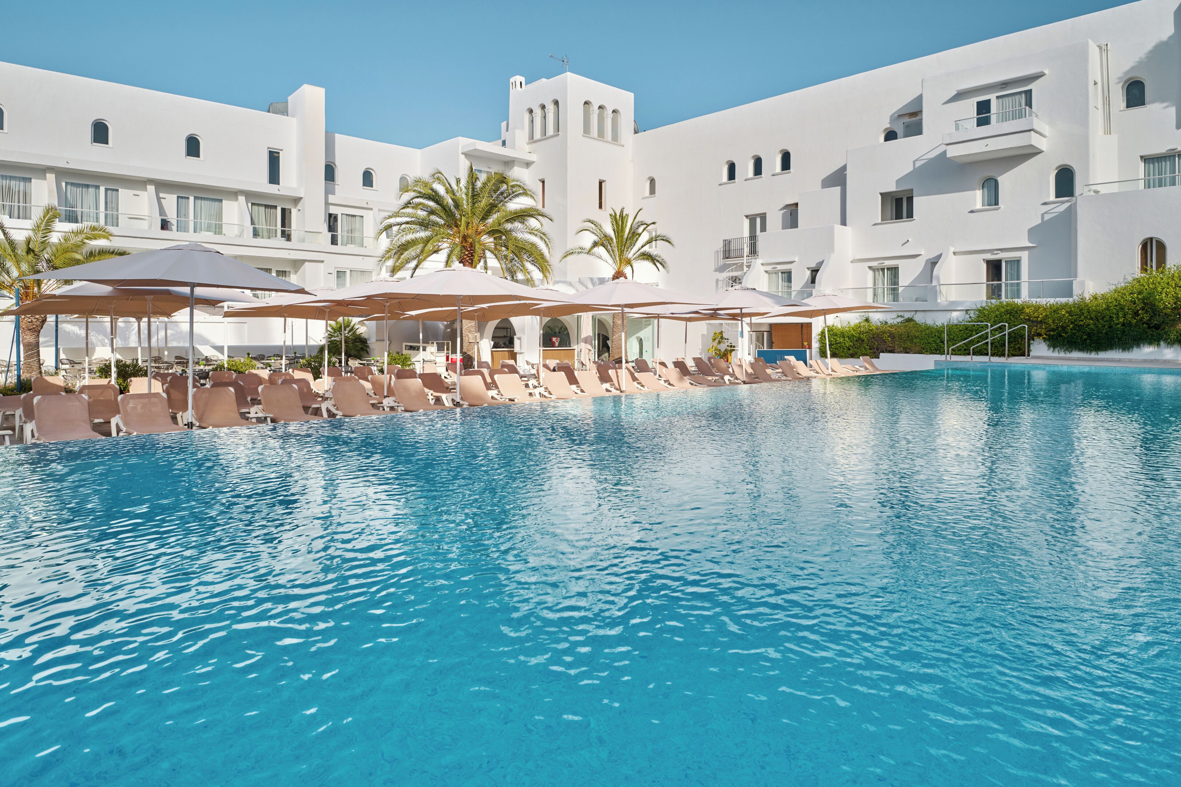 A hotel pool area with sun loungers and umbrellas, surrounded by white buildings with balconies.