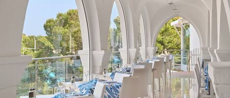 A restaurant with white arches, blue tablecloths, and a view of trees.
