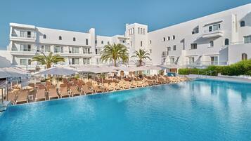 Zona junto a la piscina con sillones de descanso y sombrillas, rodeada de edificios blancos con balcones.