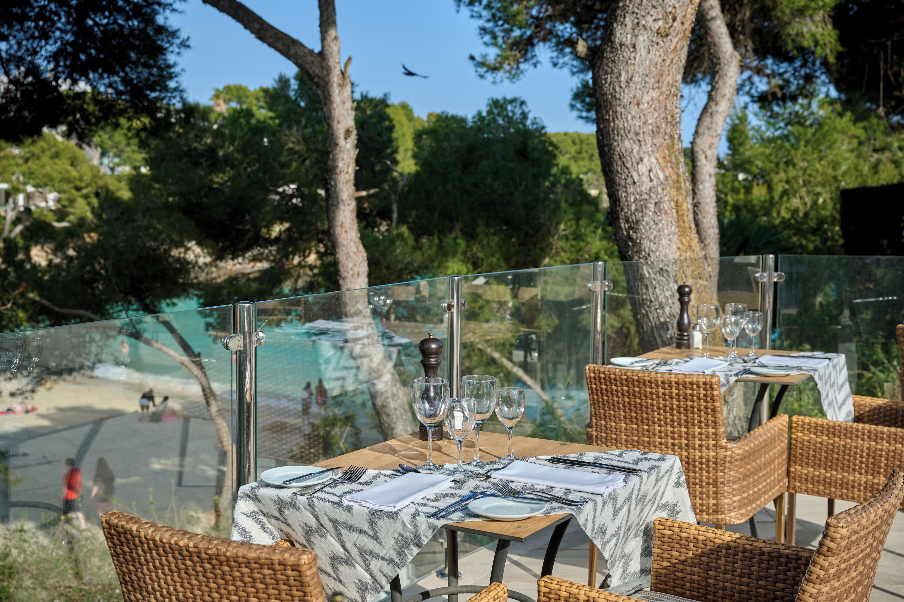 Un espacio de comedor al aire libre con mesas dispuestas para dos, con vistas a un paisaje pintoresco con árboles y una masa de agua.