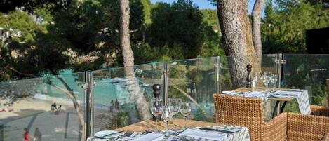 Un espacio de comedor al aire libre con mesas dispuestas para dos, con vistas a un paisaje pintoresco con árboles y una masa de agua.