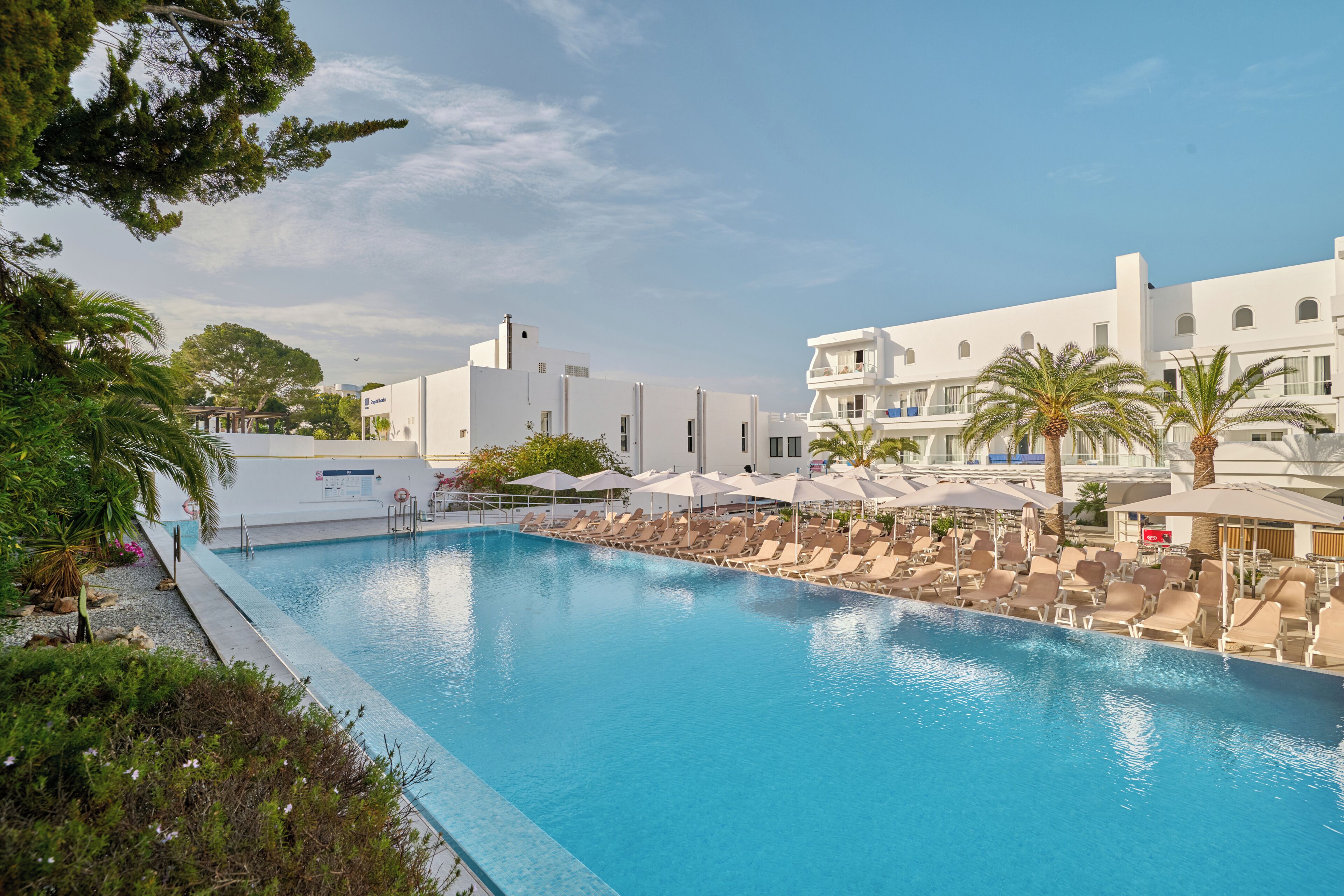 Una piscina con sillas de descanso y sombrillas, rodeada de edificios blancos y palmeras.
