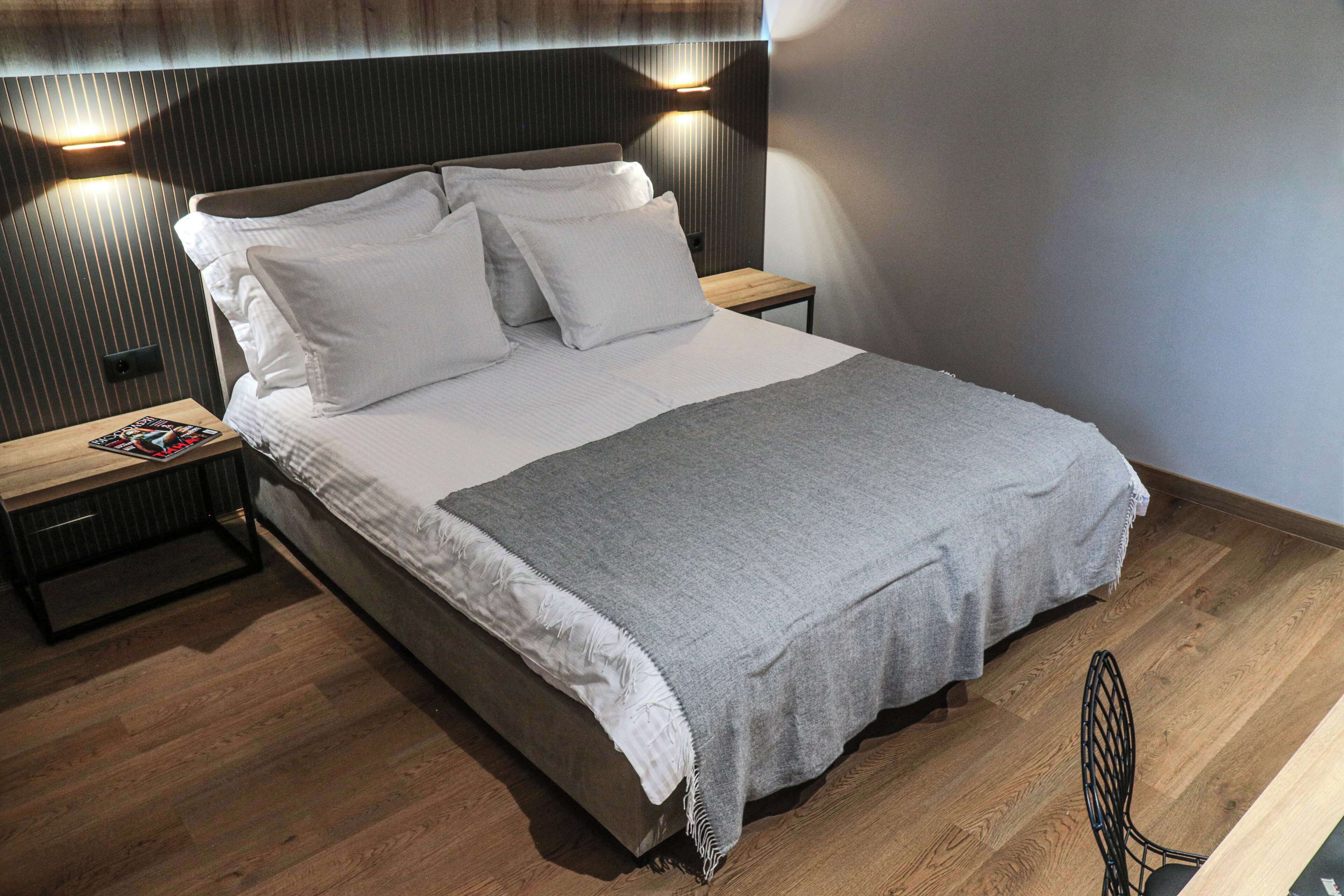 Ein ordentlich bezogenes Bett mit weißen Leinen und einer grauen Tagesdecke, ein Nachttisch aus Holz mit einer Zeitschrift und ein modernes Kopfteil mit Wandleuchten.