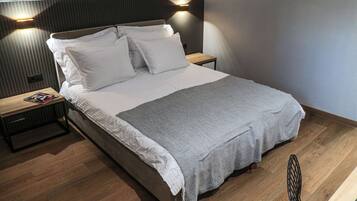 Ein ordentlich bezogenes Bett mit weißen Leinen und einer grauen Tagesdecke, ein Nachttisch aus Holz mit einer Zeitschrift und ein modernes Kopfteil mit Wandleuchten.