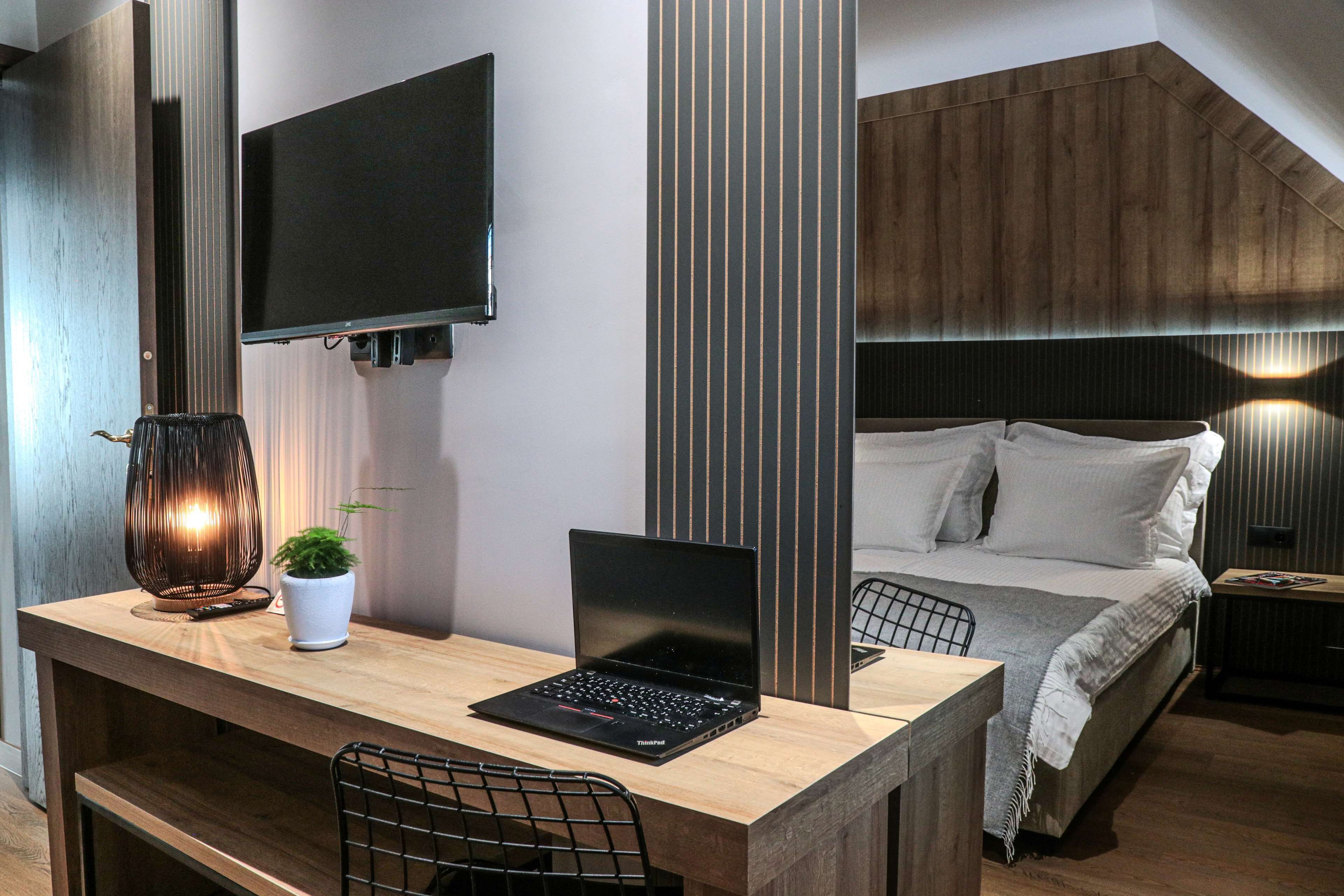 A modern hotel room with a wooden desk, a laptop, a TV, a bed with white bedding, and a small potted plant.
