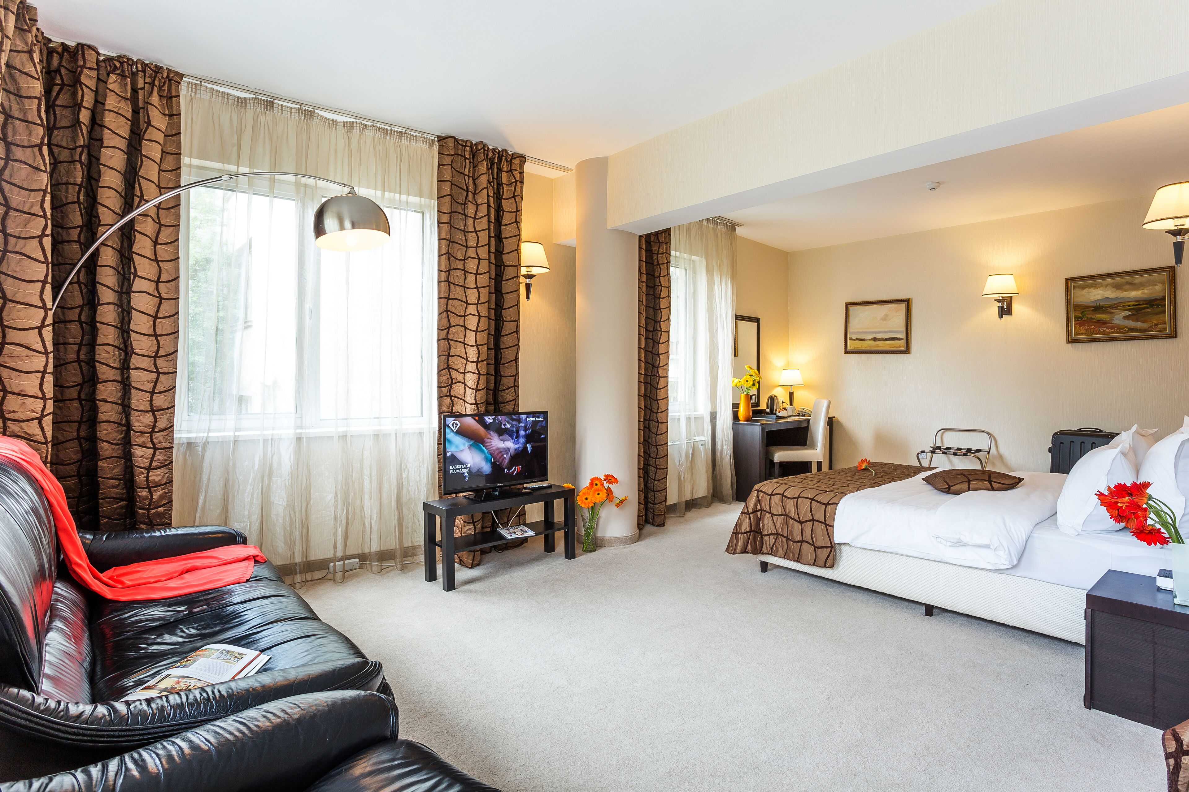 A hotel room with a leather sofa, a bed with a brown blanket, a desk with a computer, a TV, and two wall-mounted lamps.