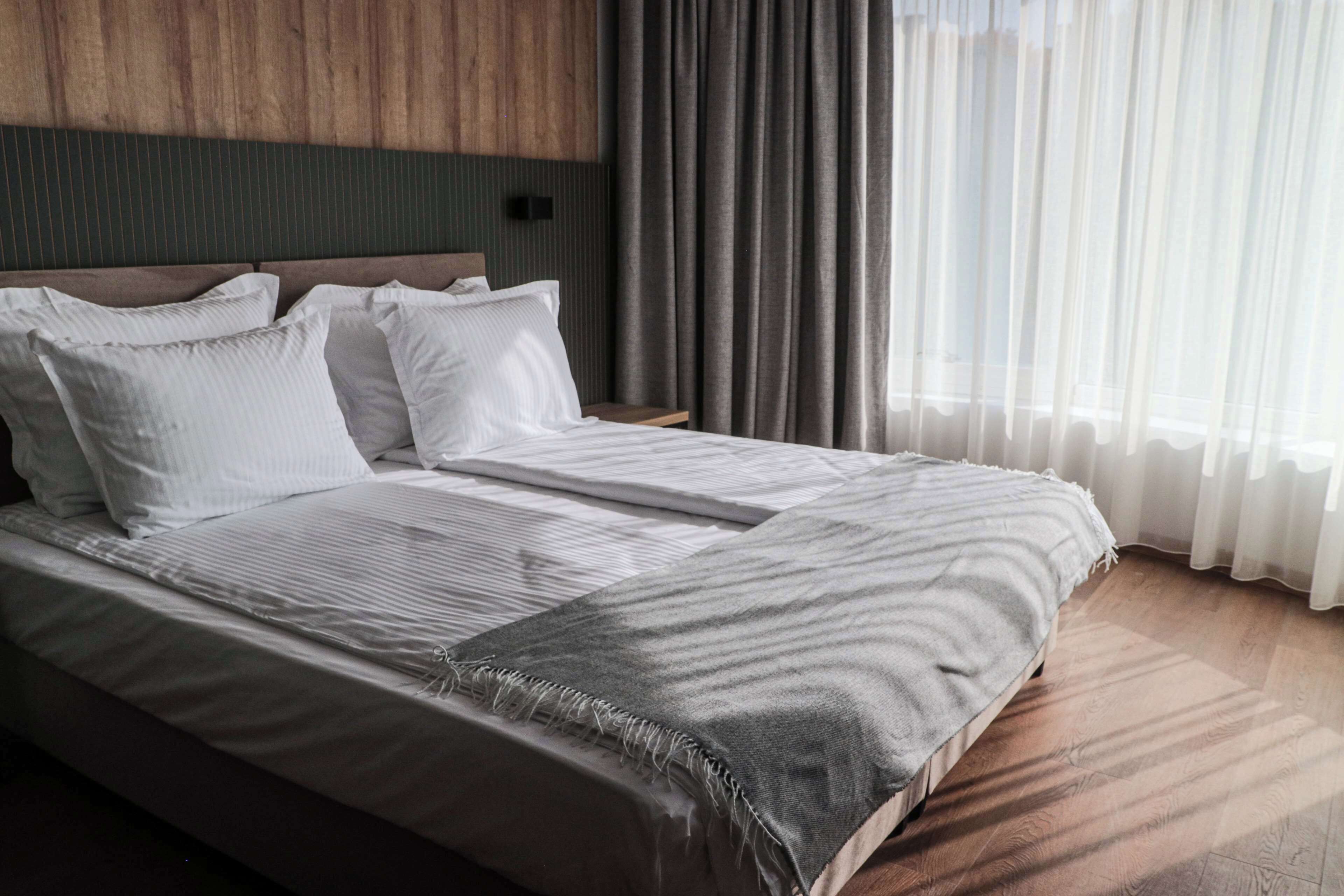A modern hotel room with a large bed, a wooden headboard, and a view of the outdoors through sheer curtains.