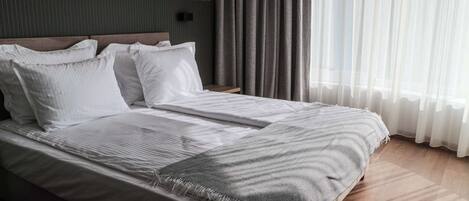 A modern hotel room with a large bed, a wooden headboard, and a view of the outdoors through sheer curtains.