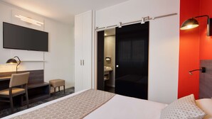 Habitación de hotel con cama, escritorio, silla, televisor montado en la pared y puerta corredera que da al baño.