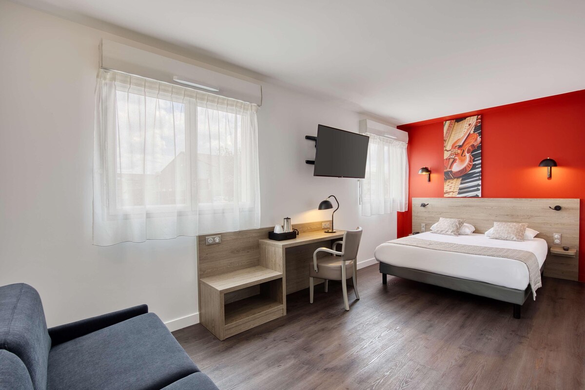 A hotel room with a bed, a desk, a chair, a TV, and a red accent wall.