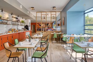 Una cafetería moderna con un interior luminoso, mesas de madera, sillas verdes y una variedad de plantas y utensilios de cocina en la barra.