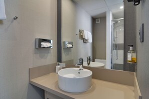 Un baño moderno con un lavabo blanco redondeado, un espejo, dispensador de toallas y cabina de ducha de vidrio.
