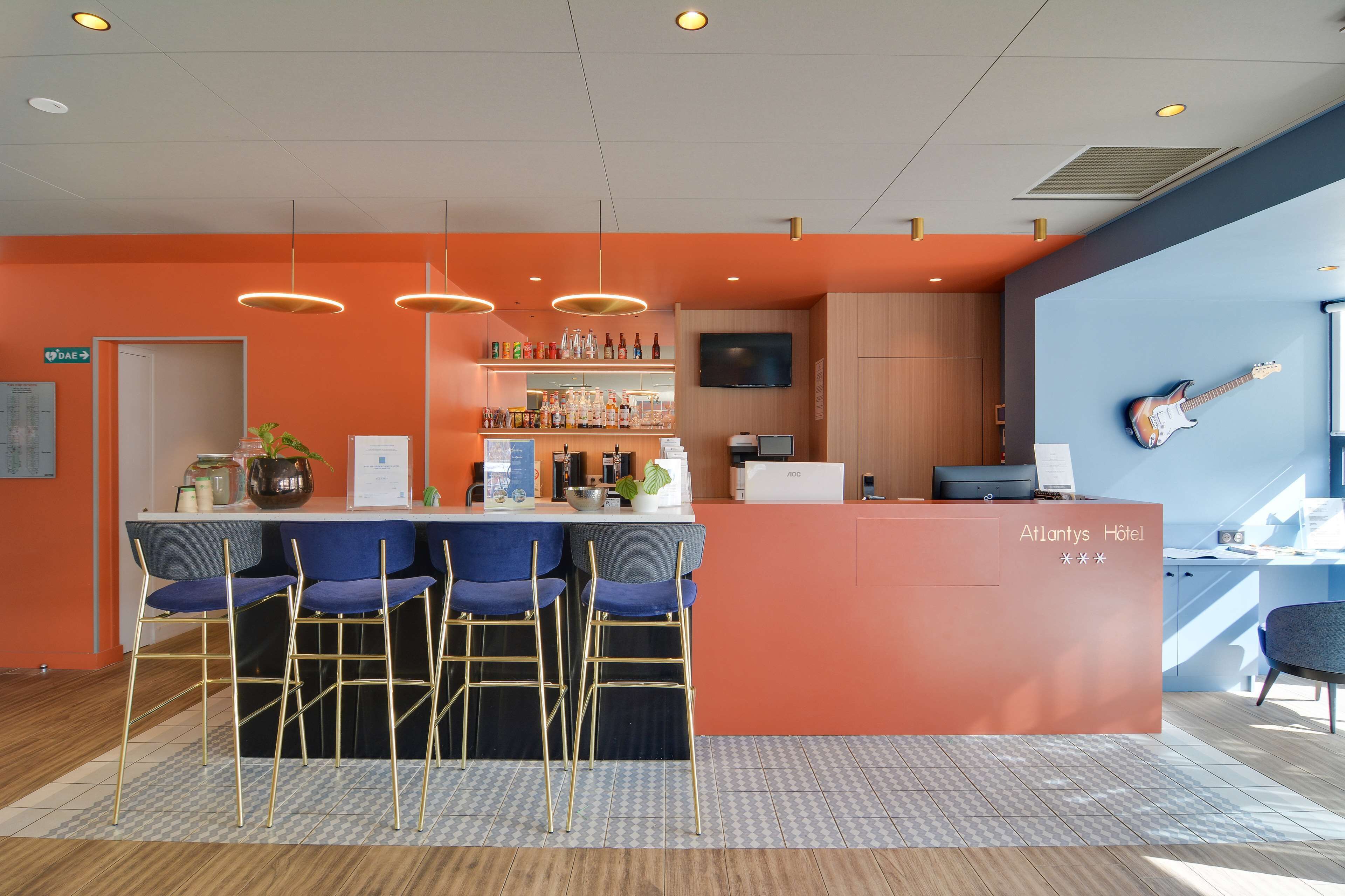 Una zona reception di un hotel con un bar, sgabelli da bar blu, una televisione e una chitarra appesa al muro.