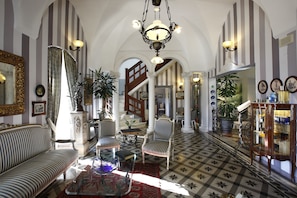 Ein geräumiges Zimmer mit gestreifter Wandgestaltung, einer Kristallleuchtre, einem Sofa, Sesseln, einem Couchtisch und einem gemusterten Boden.
